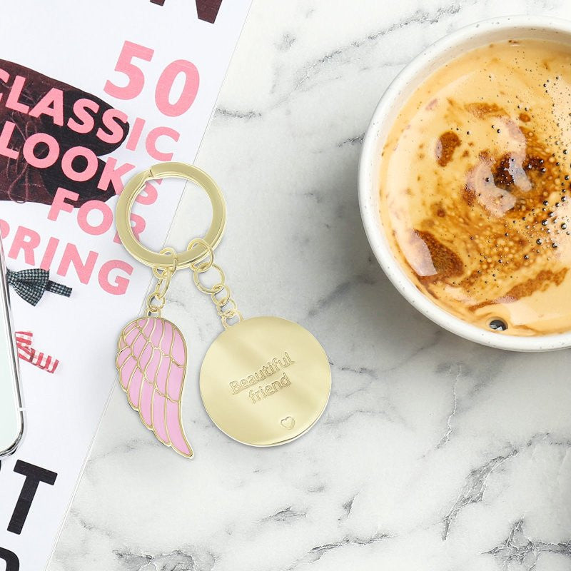 The You Are An Angel Beautiful Friend Key Chain ANGK010, featuring a pink wing charm and gold disc engraved Beautiful friend, lies on luxurious leather by a coffee cup and a magazine, celebrating lasting friendship.