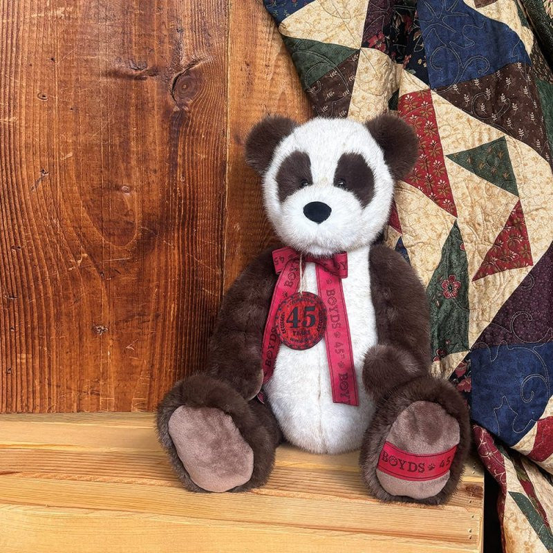The 16-inch Anni Bearsley Jointed Plush Bear by Boyds Bears (6017138) features a red ribbon around its neck and sits on a wooden bench in front of a colorful quilt and wooden wall. The ribbon displays “Boyds 45.”.