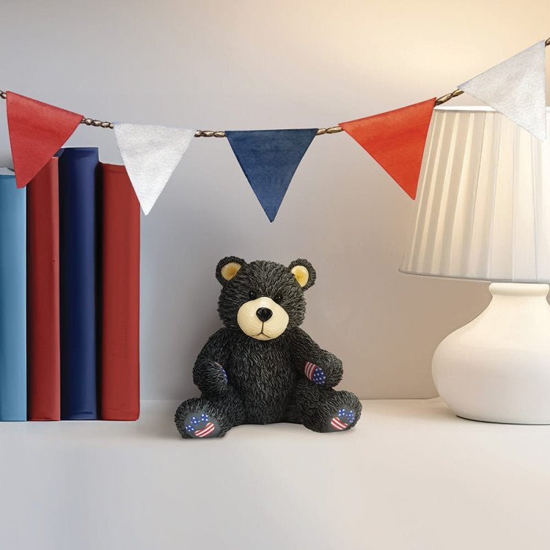 A Bubba Bearsevelt Figurine by Boyds Bears (6016928) from the 45th anniversary collection sits on a white surface beside stacked books and a lamp, with red, white, and blue bunting hanging above.