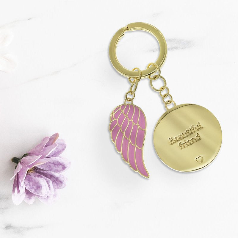 The You Are An Angel Beautiful Friend Key Chain ANGK010 features a gold keyring with a pink enamel angel wing charm and an engraved gold disc. Displayed on white marble with a light purple flower, it symbolizes lasting friendship.