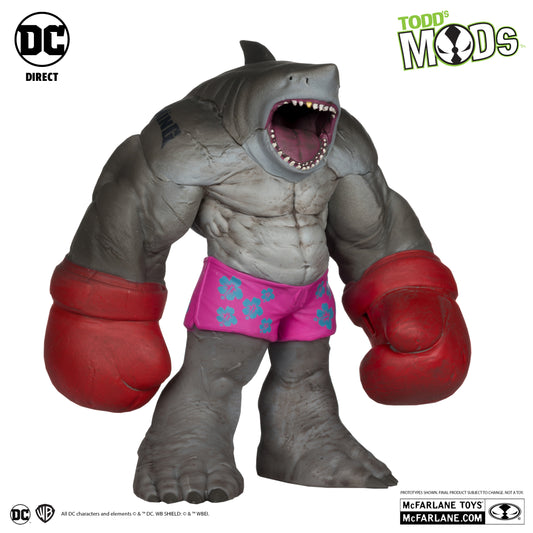 A gray, muscular King Shark figure from McFarlane Toys DC Direct Todd’s Mods wears pink shorts with blue flowers and red boxing gloves, mouth open to show teeth. Logos for Suicide Squad, DC Direct, Todd’s Mods & McFarlane Toys are in the corners.