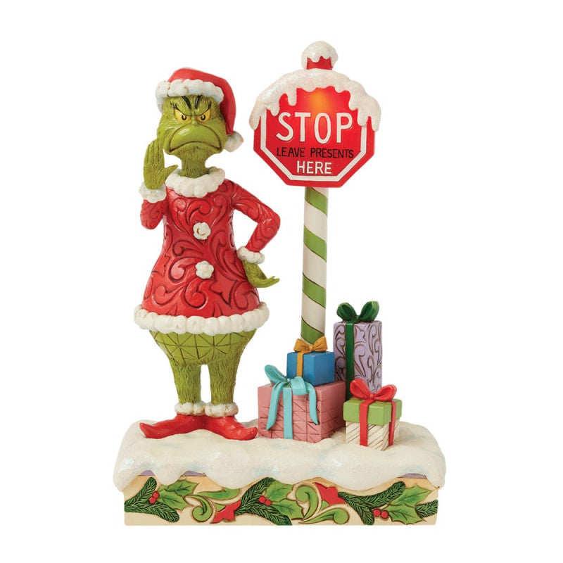 The Grinch By Jim Shore 6017063 features the Grinch in a Santa suit beside a striped pole with a flashing STOP Leave presents here sign, surrounded by gifts on a snowy, holly-trimmed base.