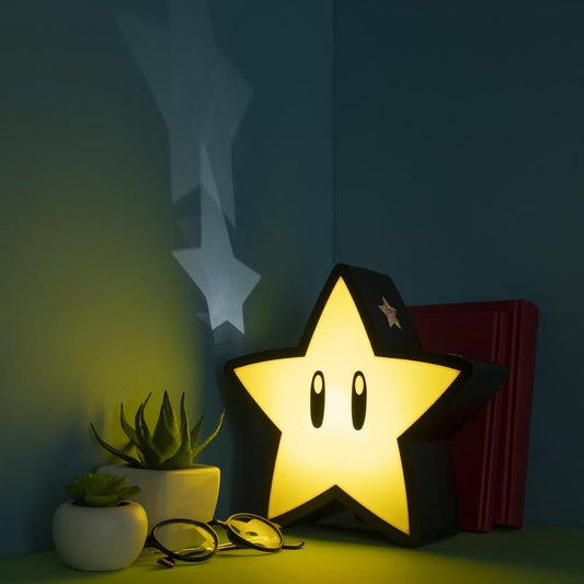 The Super Mario Star Projection Light by Paladone glows on a desk beside succulents, eyeglasses, and red books, casting a star-shaped shadow on the blue wall—an ideal collectible for fans of this iconic Nintendo product.