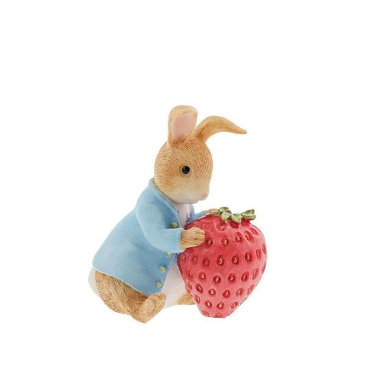 The Beatrix Potter Peter Rabbit with Strawberry Figurine from Beatrix Potter features Peter Rabbit in a blue jacket, sitting upright with his paw on a red strawberry, set against a plain white background.