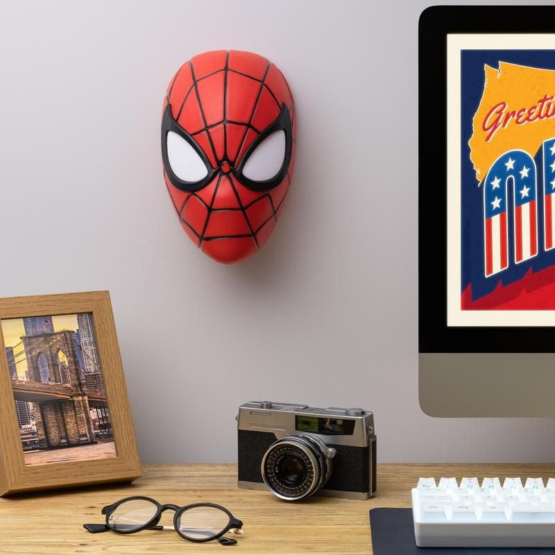 A desk holds glasses, a vintage camera, a framed photo, and a monitor. On the wall above is the Spiderman Mask Light by Paladone next to a colorful art print—a fun addition for any Marvel fan.