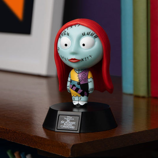 The Disney NBC Sally Icon Light by Paladone, featuring Sally with red hair, a patchwork dress, and stitches, stands on a black display base on a wooden surface near books.