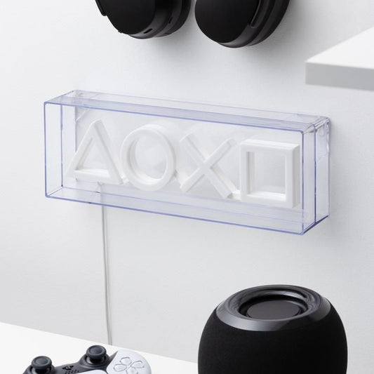 A PlayStation LED Neon Light by Paladone displays white controller icons on the wall, while a gaming console controller and black speaker rest on a white desk, creating an inviting gaming vibe with headphones hanging above.
