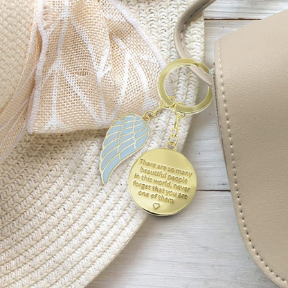 The Beautiful People Key Chain ANGK014 by You Are An Angel features a gold key chain with a blue wing charm and a round pendant engraved with “There are so many Beautiful People in this world. Never forget that you are one of them.”.