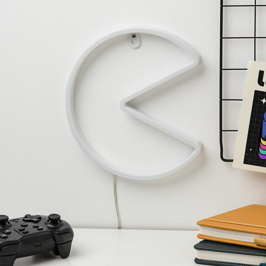 The Pac-Man Wall LED Neon Light by Paladone, shaped like PAC-MAN, is mounted above a desk with a game controller and notebooks nearby, adding the perfect retro gaming vibe for fans.