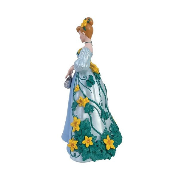 The Enesco Disney Showcase Cinderella Botanical Figurine 6015332 features Cinderella in a blue gown with green vines and large yellow flowers, holding a purse and wearing a yellow flower in her hair, viewed from the side.