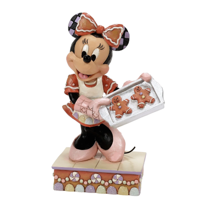 The Disney Traditions Baking Bliss (Minnie Gingerbread Cookies) Figurine by Jim Shore 6016881 features Minnie Mouse in a pink bow and apron holding a tray of gingerbread cookies, on a candy-themed base. Pre-order now from Disney Traditions.