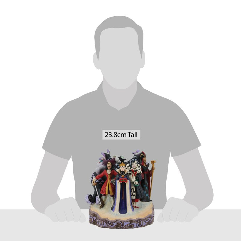 Seated at a table, a person admires the 23.8 cm tall Enesco Disney Traditions Villains Carved by Heart By Jim Shore 6010880 figurine featuring Maleficent, Cruella de Vil, the Evil Queen, and Jafar.