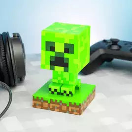 The Minecraft Creeper Icon Light by Paladone, featuring the iconic green pixelated design on a square base, sits on a desk beside black headphones and a game controller.
