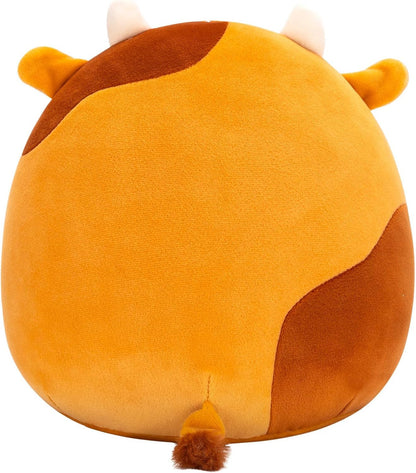 The Squishmallows Jericho the Gingerbread Cow collectible plush is mostly orange with brown patches, small white horns, orange ears, and a tiny brown tail. Seen from the back, it has a round, plush cow shape. Brand: Squishmallows.