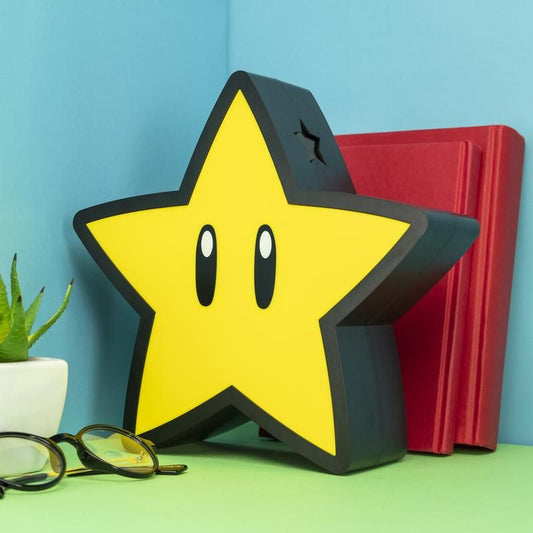 A yellow Super Mario Star Projection Light by Paladone with black eyes sits on a green surface beside a red book, a small potted plant, and glasses, all set against a blue wall.