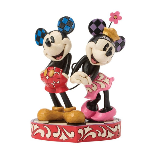 The Disney Traditions Tails of True Love Mickey & Minnie Love Figurine by Jim Shore features Mickey in red shorts and blue checkered ears, and Minnie in a pink skirt, flower hat, and purple checkered ears, standing together holding hands.