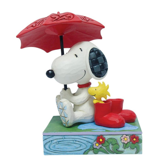 The Snoopy Under Umbrella Figurine by Peanuts By Jim Shore features Snoopy and Woodstock relaxing under a red umbrella beside rain boots, on a decorative base with blue puddles and flowers.