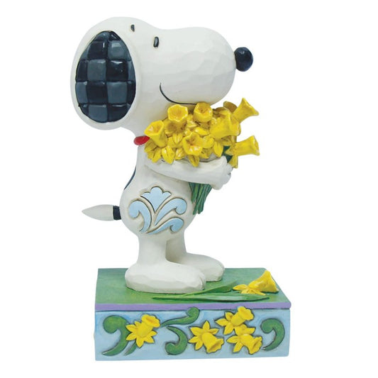 The Snoopy with Daffodils Figurine by Peanuts By Jim Shore features Snoopy smiling and holding a bouquet of yellow daffodils, standing on a floral patterned base with checkered ears.