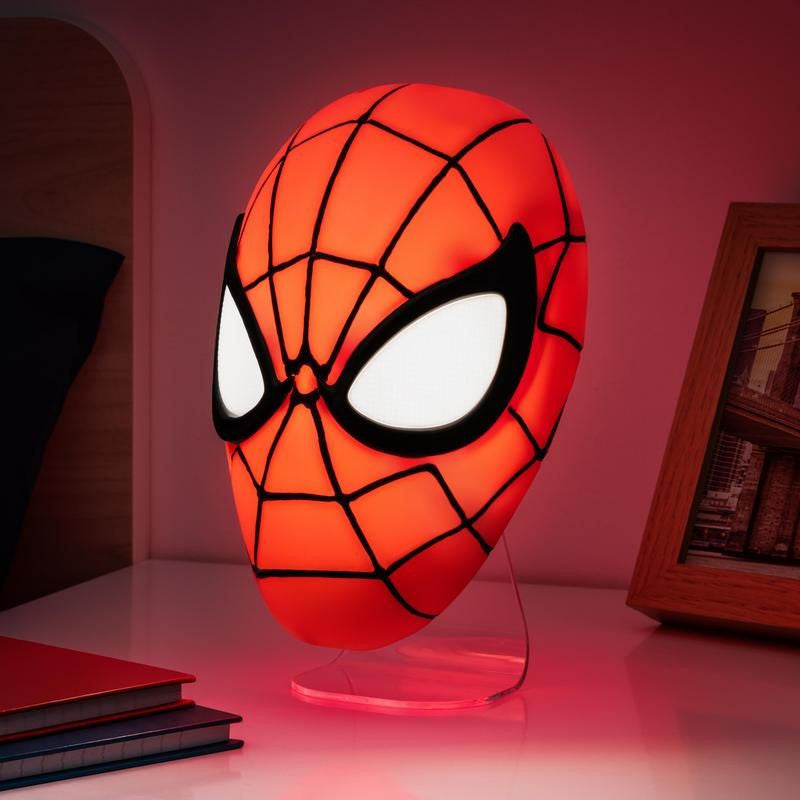 The Spiderman Mask Light by Paladone glows red and white on a white table beside a photo frame and closed notebooks, illuminating the dim room—an ideal decor piece for Marvel fans or anyone who loves Spiderman.