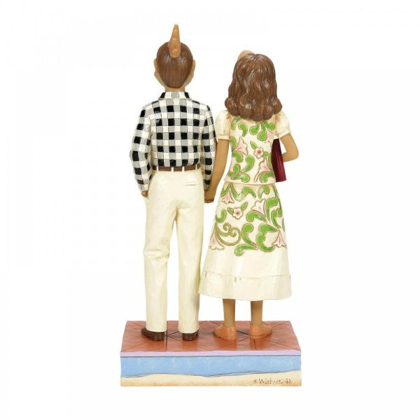 The Beetlejuice By Jim Shore Adam & Barbara Maitland Figurine (6016176) shows the couple standing side by side, with Adam in a black-and-white checkered shirt and Barbara in a white dress with green floral patterns holding a red book.