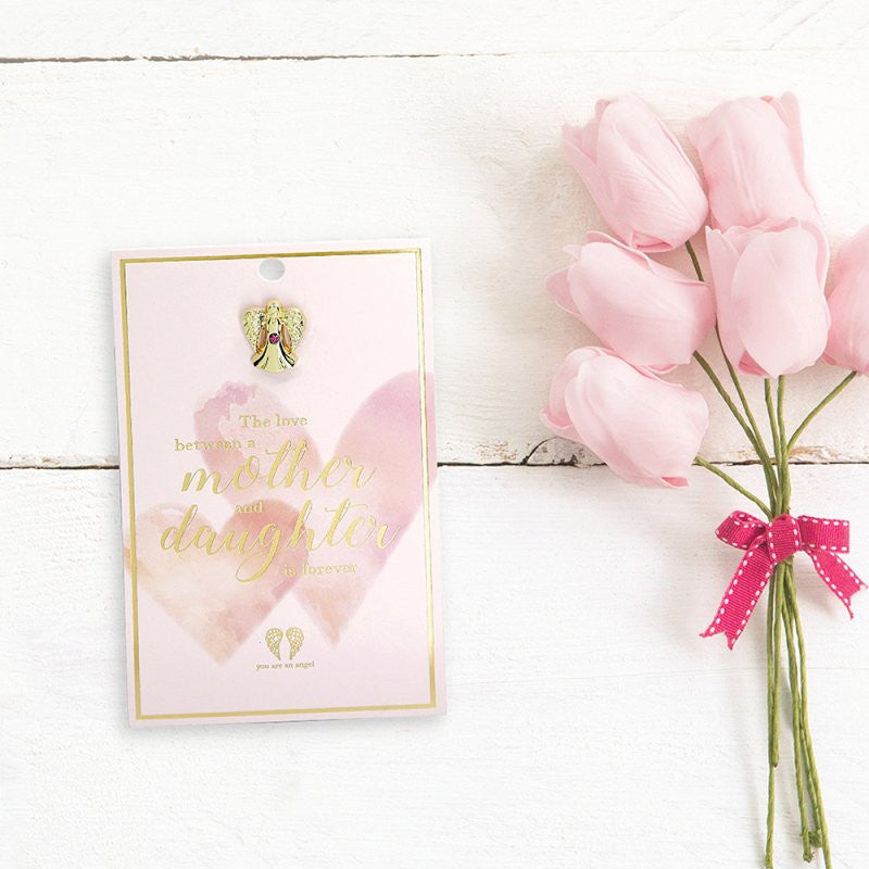 The You Are An Angel Mother And Daughter Pin Card ANCP084, featuring the words The love between a mother and daughter is forever, rests beside pink tulips with a polka-dot ribbon on a white wooden surface.