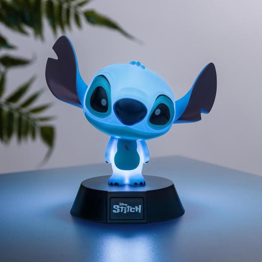 The Disney Stitch Icon Light by Paladone features a blue night light figure on a round black base with the Stitch logo, perfect for Disney fans and collectors. Set on a light blue surface with blurred plant leaves in the background.