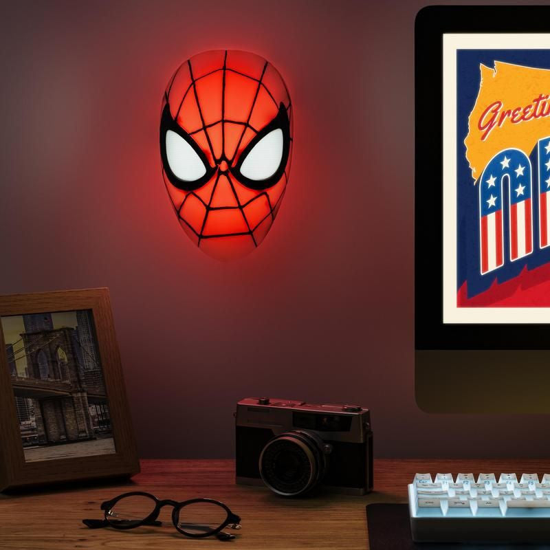 The Spiderman Mask Light by Paladone is mounted above a desk with a photo frame, camera, glasses, monitor, and keyboard—ideal Marvel-themed decor for fans of the superhero.