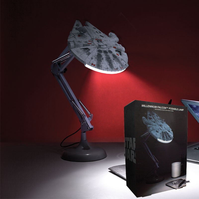 The Star Wars Millennium Falcon Posable Desk Light V3 by Paladone is a licensed lamp modeled after the iconic ship, casting light on your desk. Its box, featuring the lamp, sits nearby against a dark red background.