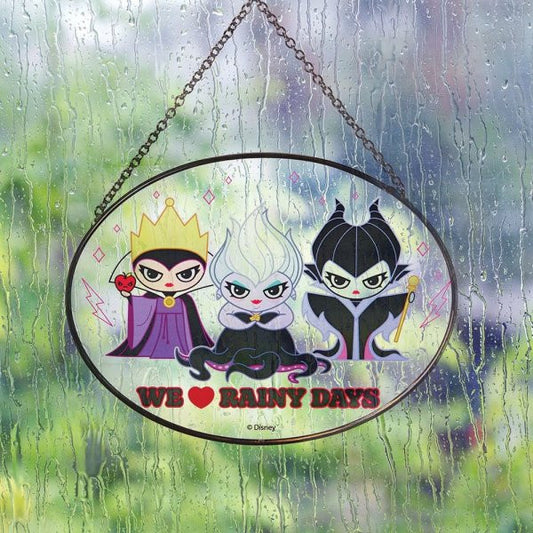 The Villains Suncatcher by Disney Garden 6014572, featuring three cartoon Disney Villains, hangs on a rainy window. A sign with We ♥ Rainy Days captures the moody scene.