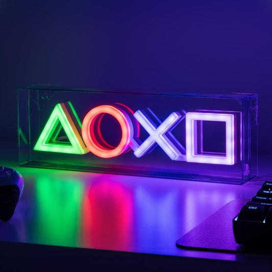 The PlayStation LED Neon Light by Paladone glows with green, red, blue, and purple PlayStation controller icons, creating the ideal gaming atmosphere on your desk, with a keyboard and controller nearby.