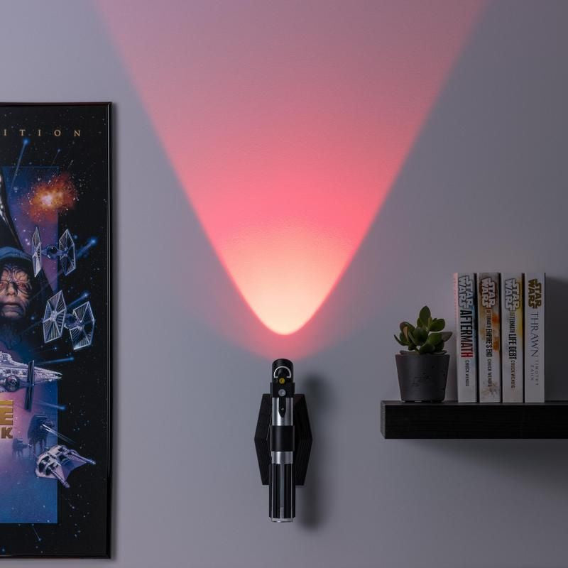 The Paladone Star Wars Lightsaber Uplighter, an officially licensed Darth Vader-inspired product, mounts on the wall and emits a red upward beam. Next to it are a framed poster and a shelf with Star Wars books and a small plant.
