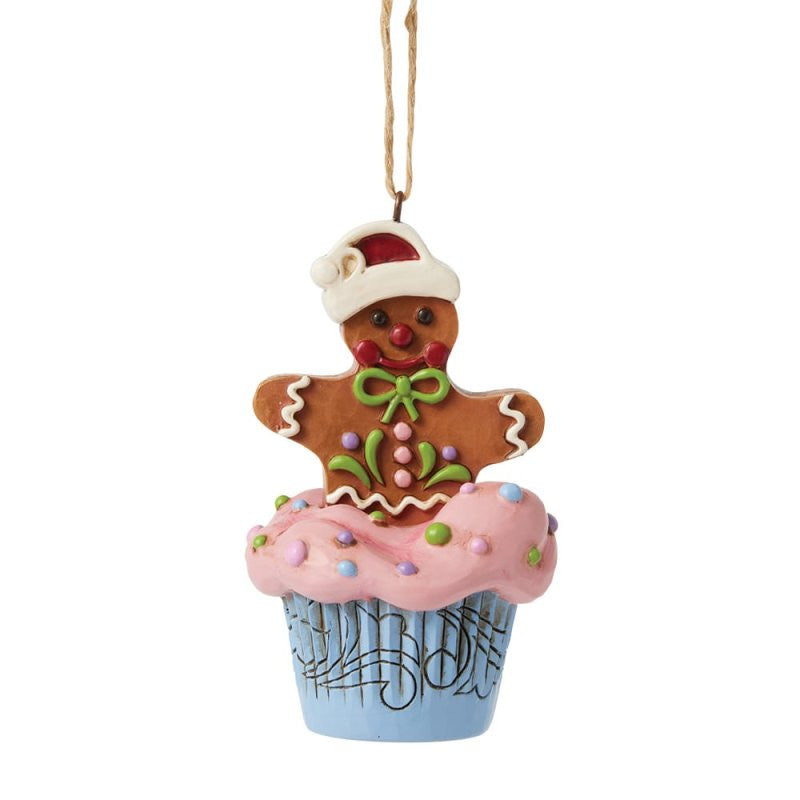 The Heartwood Creek By Jim Shore Gingerbread Cupcake Hanging Ornament 6017271 features a cheerful gingerbread man in a Santa hat and green bow sitting on a blue cupcake with pink frosting and sprinkles, hung by a brown string.