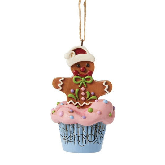 The Heartwood Creek By Jim Shore Gingerbread Cupcake Hanging Ornament 6017271 features a cheerful gingerbread man in a Santa hat and green bow sitting on a blue cupcake with pink frosting and sprinkles, hung by a brown string.
