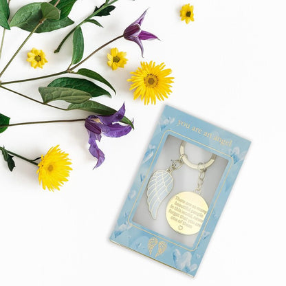 The Beautiful People Key Chain ANGK014 by You Are An Angel, featuring an angel wing charm and engraved tag, rests in blue You Are An Angel packaging. Yellow and purple flowers surround it on a white background.