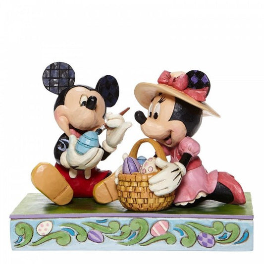 Disney Traditions Mickey and Minnie Mouse sit together. Mickey paints an Easter Egg and Minnie holds the basket figurine