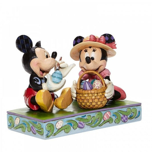 Disney Traditions Mickey and Minnie Mouse sit together. Mickey paints an Easter Egg and Minnie holds the basket figurine