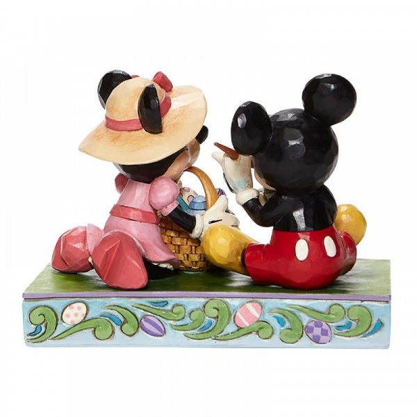 Disney Traditions Mickey and Minnie Mouse sit together. Mickey paints an Easter Egg and Minnie holds the basket figurine