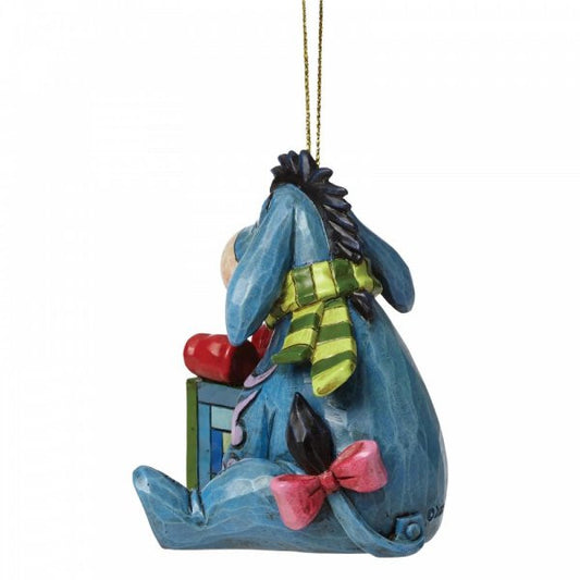 The Disney Traditions Eeyore Hanging Ornament A27553 features Eeyore with a pink bow and green striped scarf beside a gift box with a red heart, hanging by a gold string—an adorable piece from Disney Traditions.