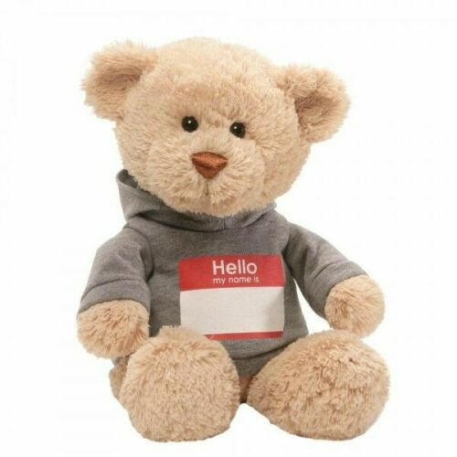 The GUND Hello My Name Is... Bear Plush by GUND is a light brown teddy bear in a grey hoody with a red and white name tag, sitting upright against a plain white background.
