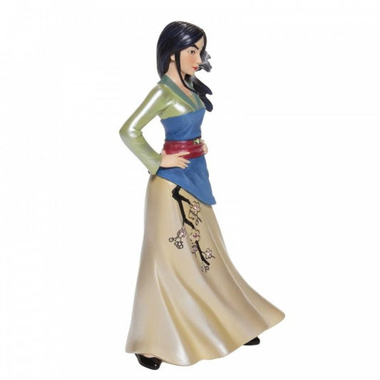 The Enesco Disney Showcase Mulan Couture de Force Figurine 6007187 features Mulan with long dark hair, a green top, blue sash, red belt, and cream skirt adorned with cherry blossoms, standing confidently hands on hips.