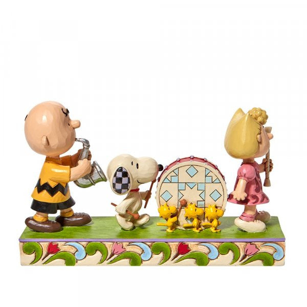 The Peanuts Parade Figurine By Jim Shore features Charlie Brown, Snoopy, and Sally in a parade with classic Jim Shore detailing—Snoopy plays a drum, Charlie Brown has a horn, Sally holds a baton, and three yellow birds lead the way.