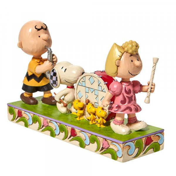 The Peanuts Parade Figurine by Jim Shore depicts Charlie Brown with a whistle, Sally with a baton, and Snoopy drumming alongside three Woodstocks on a patterned base, all with signature Peanuts By Jim Shore detailing.