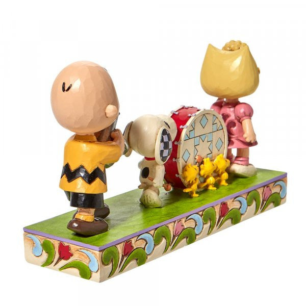 The Peanuts Parade Figurine by Jim Shore features Charlie Brown with a bat, Snoopy with a bass drum, Sally in pink, and two yellow birds on a decorative base with classic Jim Shore detailing. Brand: Peanuts By Jim Shore.