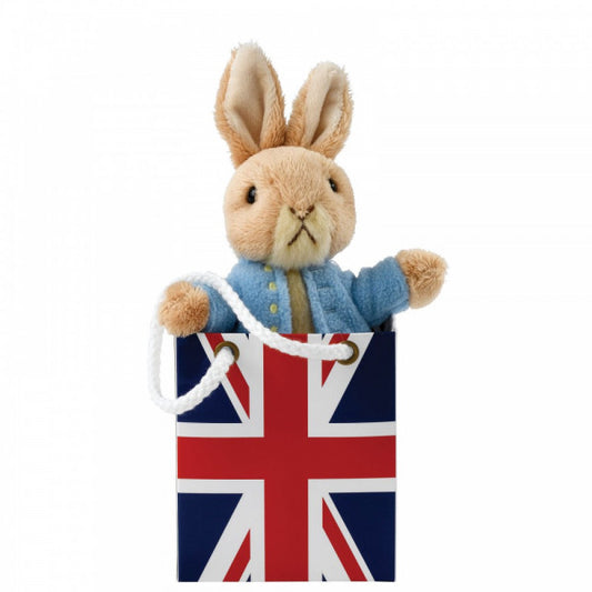 The GUND Beatrix Potter Peter Rabbit In Union Jack Bag Plush features Peter Rabbit, wearing a blue jacket, sitting inside a gift bag adorned with the United Kingdom flag.