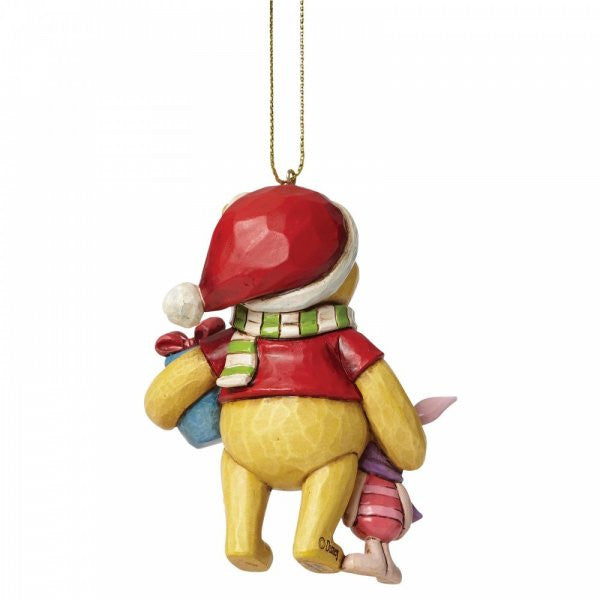 The Disney Traditions Winnie the Pooh and Piglet Hanging Ornament (A27551) shows Pooh in a Santa hat and striped scarf, holding Piglet’s hand and a blue sack, both seen from behind. It hangs from a gold string.