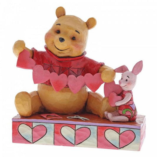 Disney Traditions Piglet and Winnie the Pooh sit close beside each other as they trace and snip a handmade garland of hearts figurine