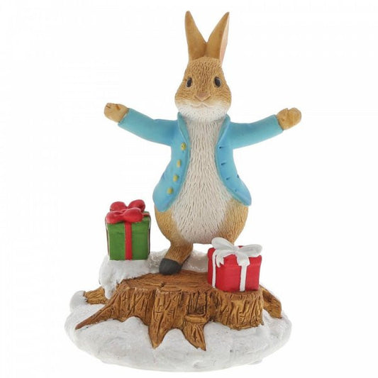 The Beatrix Potter Peter Rabbit With Presents Figurine features Peter Rabbit in a blue jacket standing on a snowy stump with arms open, beside green and red wrapped gifts—a delightful tribute to the classic tales.