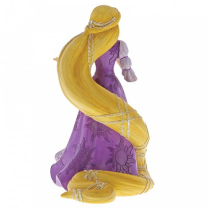 The Disney Showcase Rapunzel Figurine 6001661 features Rapunzel with long braided blonde hair adorned with white flowers, in a purple dress with sun patterns. The cast stone figure, by Disney Showcase, is shown from the back.