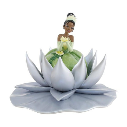 The Enesco Disney 100 Disney Showcase Tiana Icon Figurine (6013335) features Princess Tiana in a green dress and tiara, sitting in a silver lotus flower and holding a yellow blossom—ideal for celebrating Disney 100.