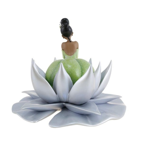 The Enesco Disney 100 Disney Showcase Tiana Icon Figurine (6013335) features Tiana with a dark bun and sparkly green dress, sitting with her back to the viewer on a silver lotus flower—a delightful collectible.